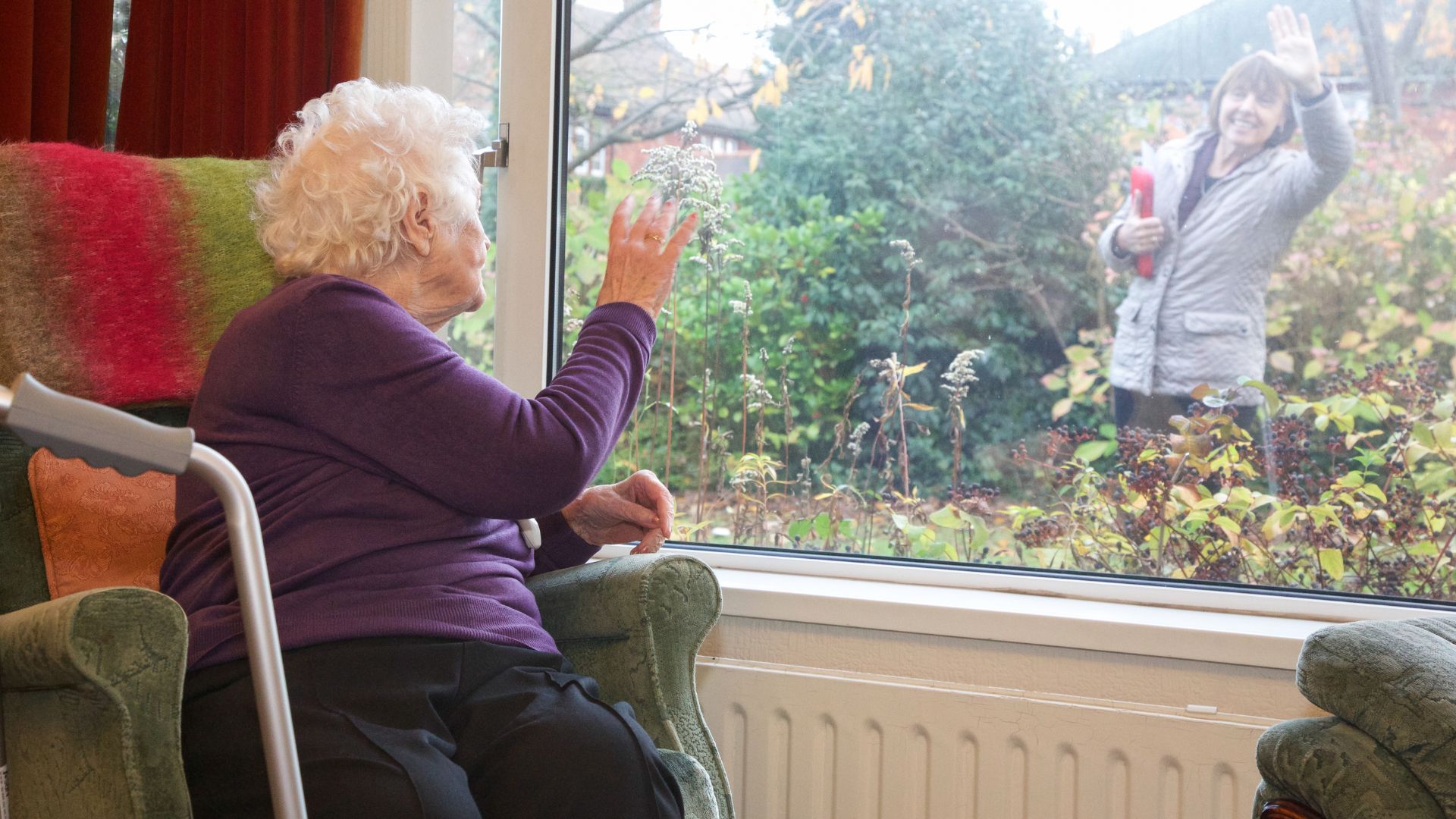Senior woman in a skilled nursing facility waving through the window to a visiting family member outside.