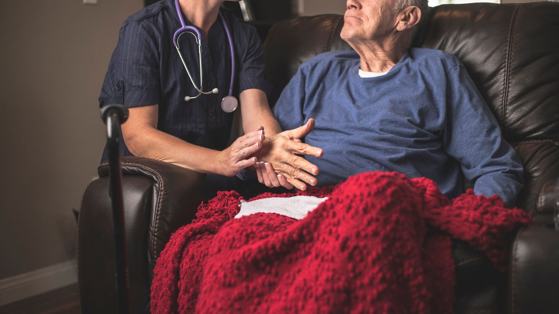 Nurse providing hospice care, holding the hands of an elderly man sitting in a recliner with a red blanket, offering comfort and compassion at home.